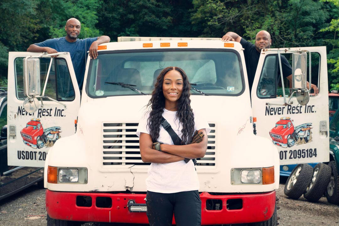 slender African American woman with long black hair and arms folded standing in front of large white truck with doors open. Two men are standing behind truck doors