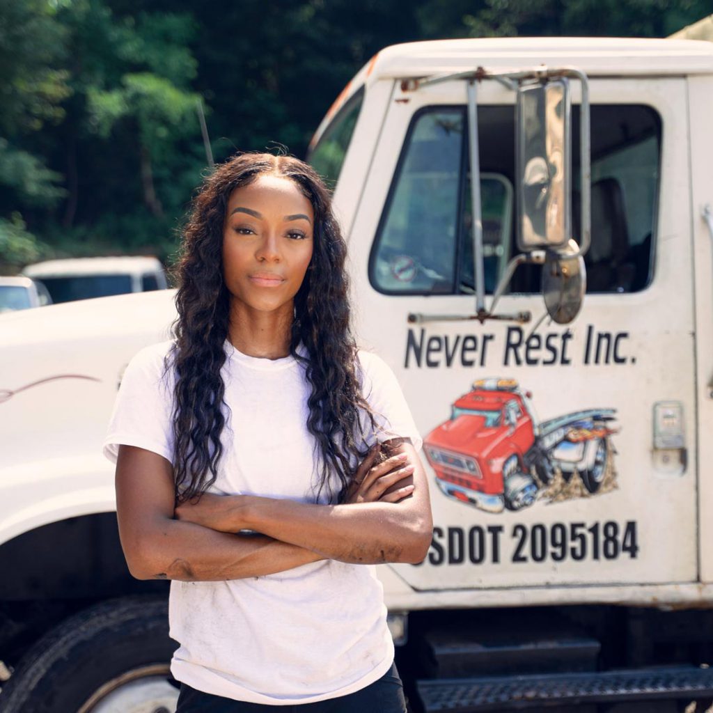 slender African American woman with long black hair and arms folded standing in front of large white truck with company name on the door